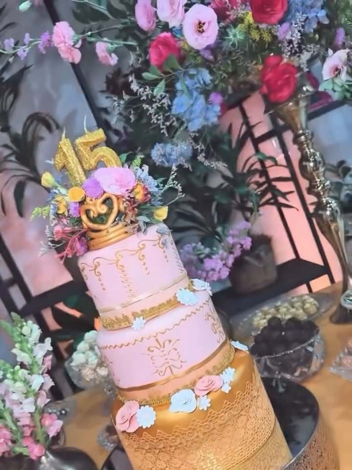 Bolo de 15 anos rosa e dourado com topo de flores, cercado de arranjos coloridos