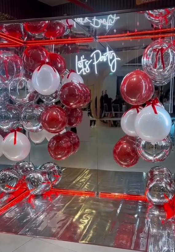 Cabine espelhada decorada com balões vermelhos, brancos e prateados e letreiro neon