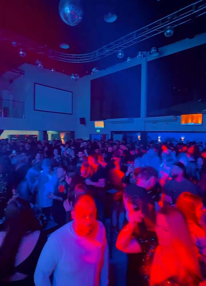 Salão lotado durante a festa, com luzes coloridas e globo espelhado