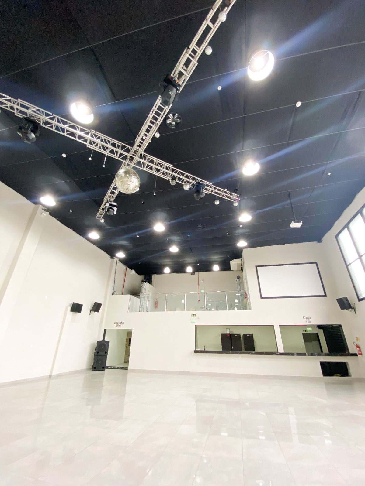 Salão principal visto da entrada: piso claro, teto preto com treliças de iluminação e o mezanino envidraçado ao fundo